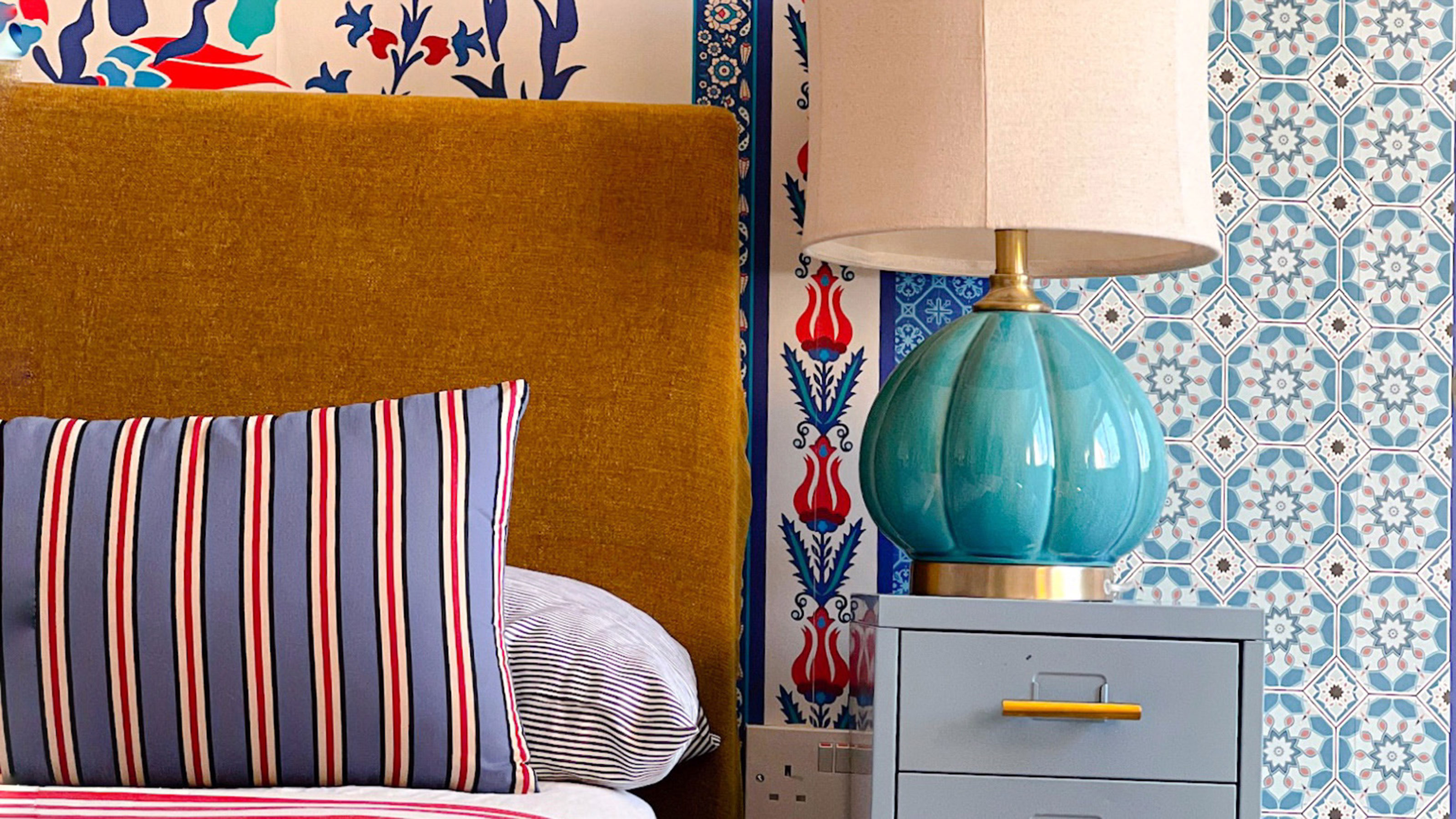 Bedroom with ochre upholstered headboard, patterned walls, pattern mix and matched pillow and bed covering beside pale blue-green storage unit with turquoise bedside lamp