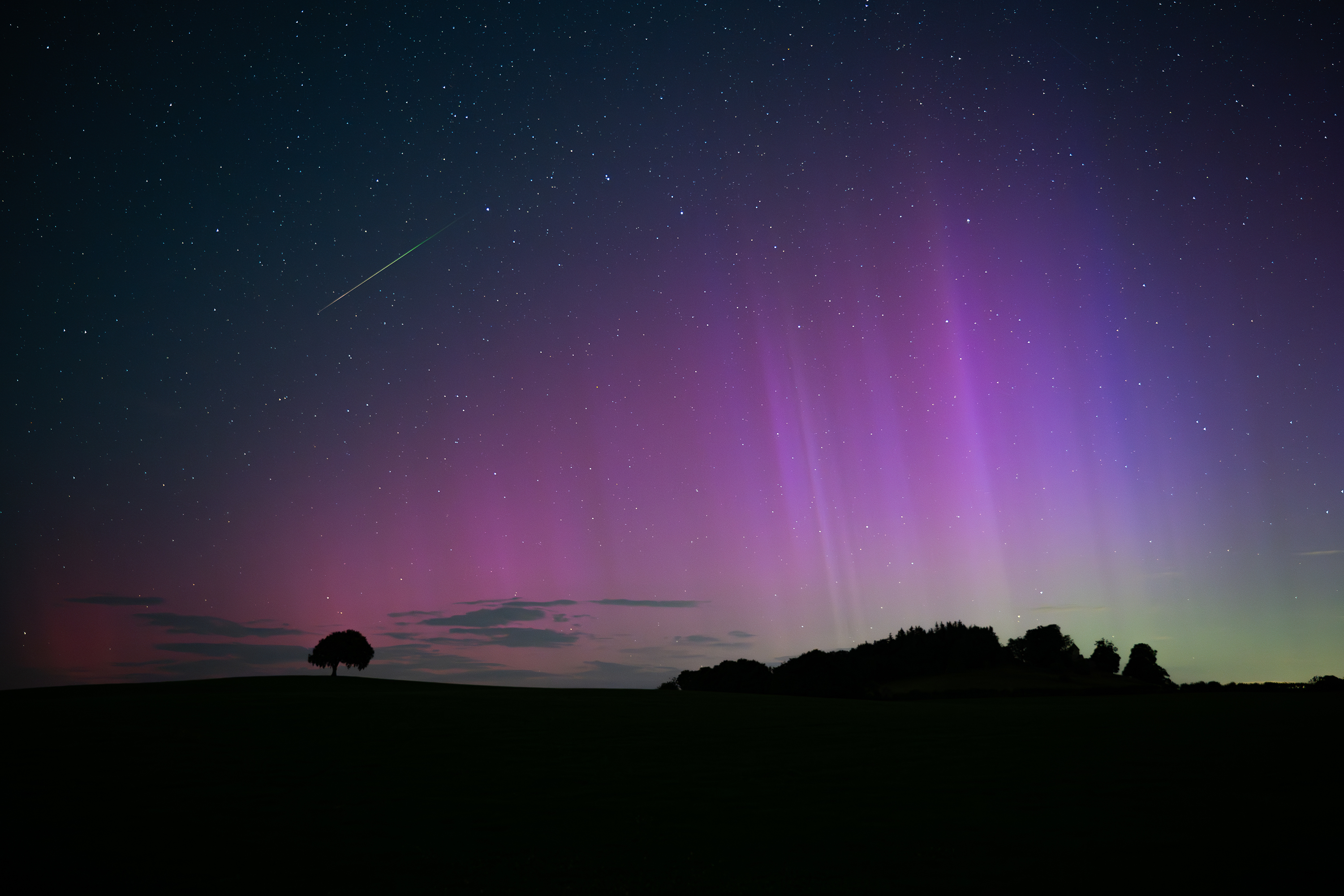 Cortinas de luz violeta y azul llenan el cielo con la larga cola de una estrella fugaz en la esquina superior izquierda de la imagen.