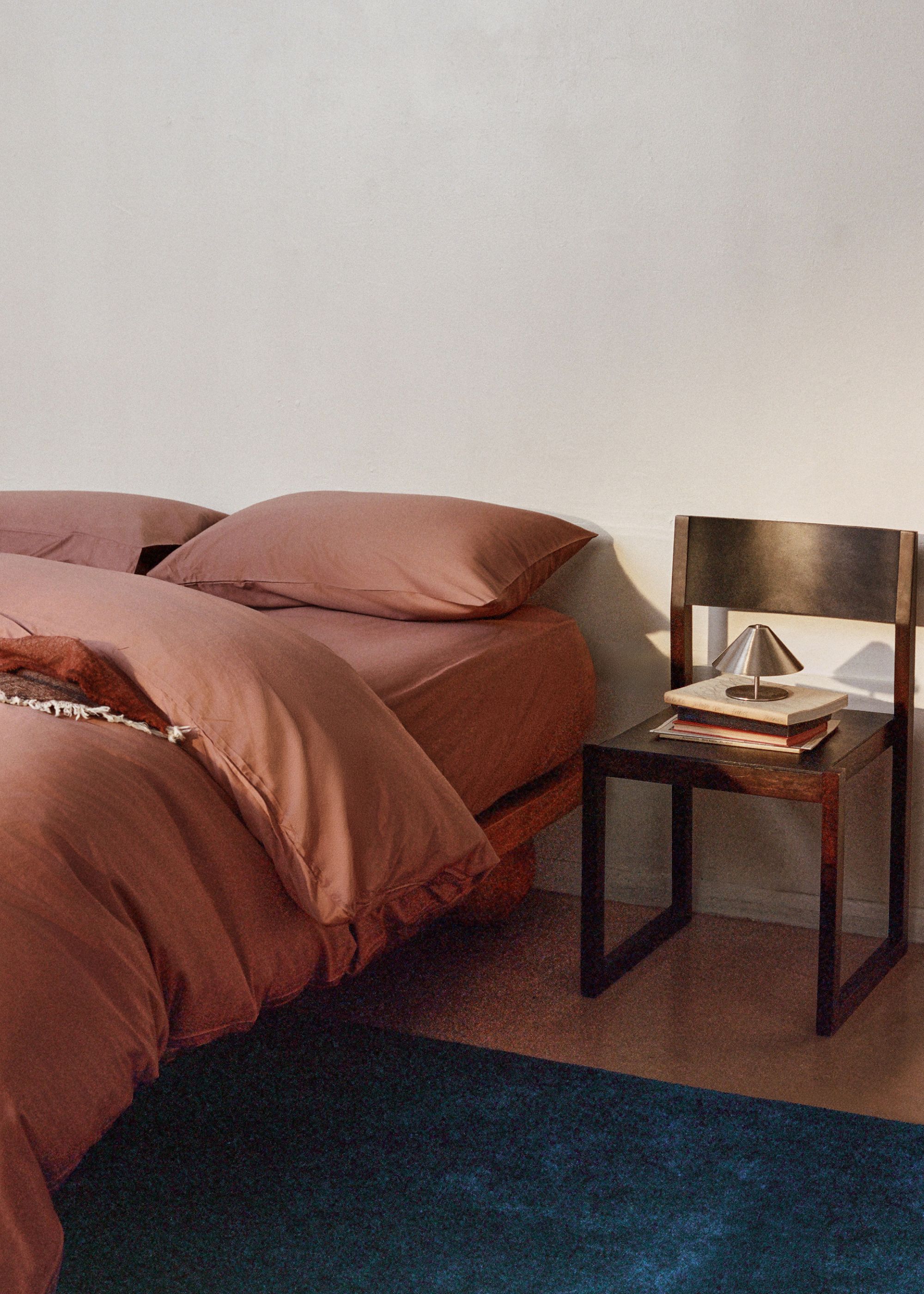 A bedroom with a chair as a side table with a stack of books and a short chrome lamp beside a bed with clay-colored bedding