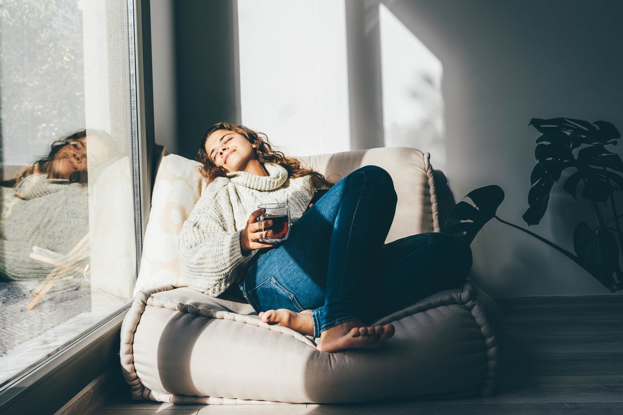 A woman relaxing in a comfy chair, with a warm tea, with the sun on her face through a nearby window.