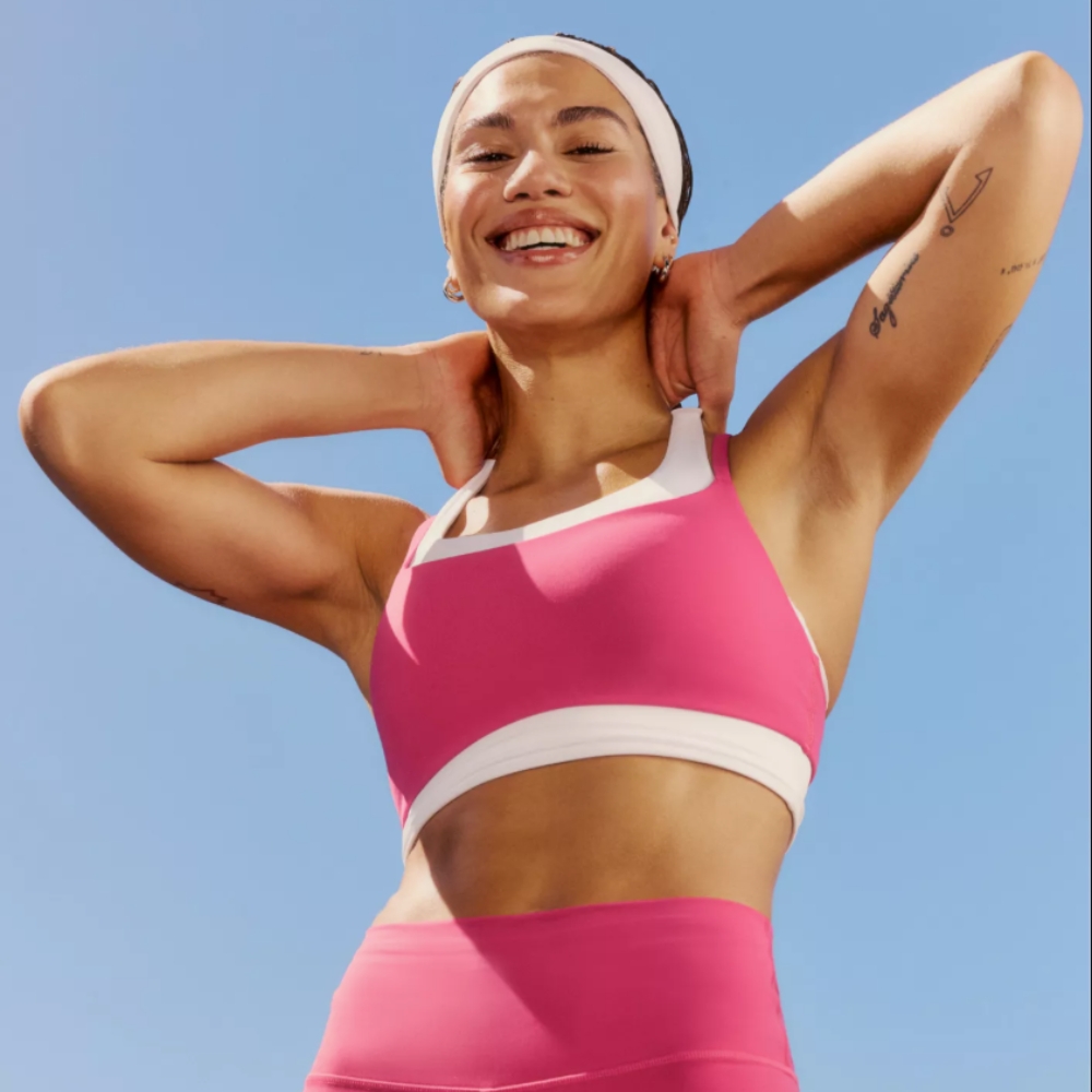 Woman in pink and white Alo Yoga set stretching her arms, with blue sky behind her