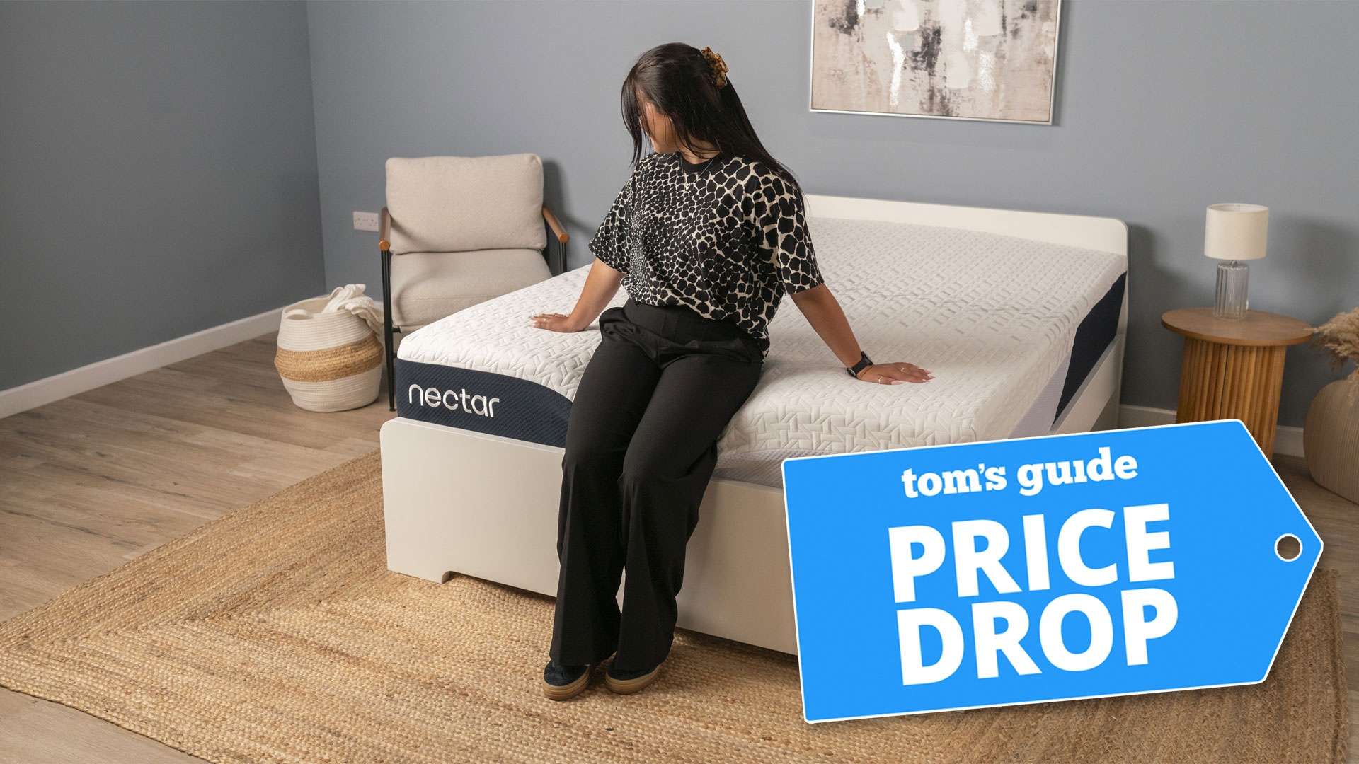 A woman sitting on the end of the Nectar Classic mattress in a bedroom, with a Tom's Guide 'PRICE DROP' badge overlaid