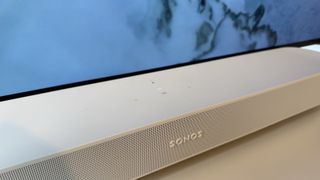 A white Sonos Beam Gen 2 soundbar photographed on a white desk in front of an OLED TV