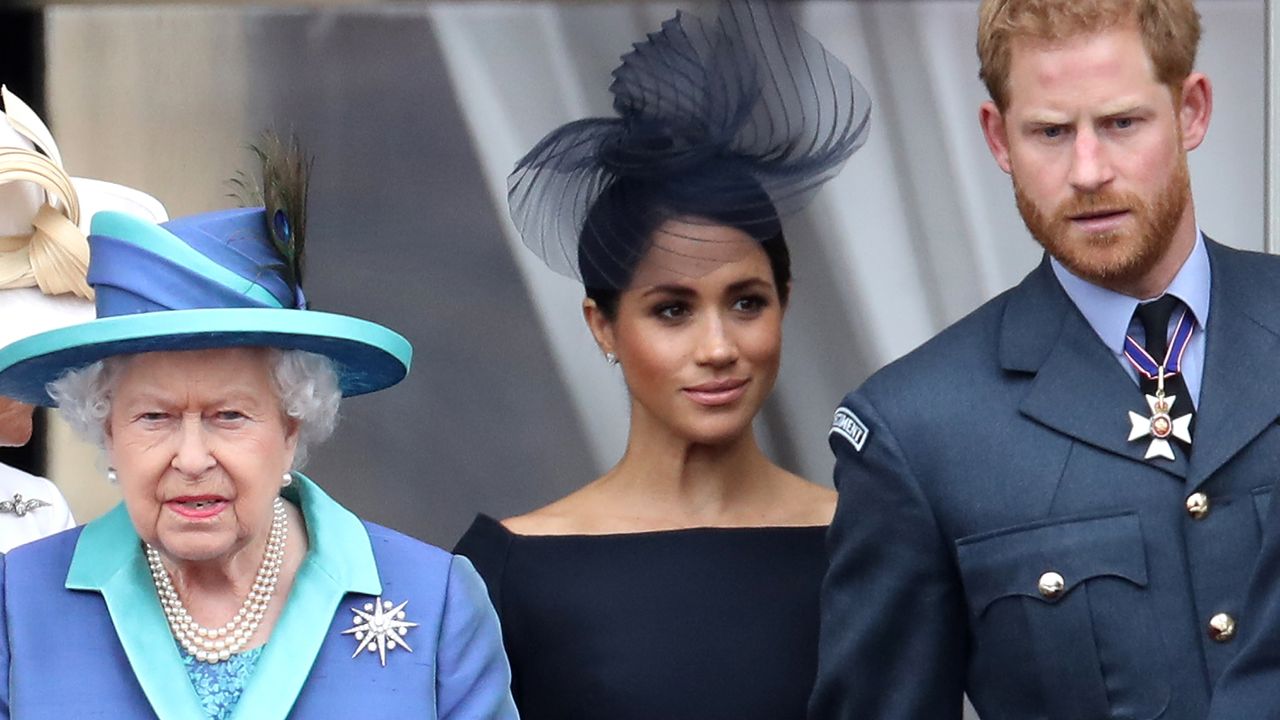 Queen Elizabeth wears a blue and turquoise coat and matching hat as she stands on the Buckingham Palace balcony with Meghan Markle, the Duchess of Sussex, who is wearing a black outfit, and Prince Harry, who is wearing military uniform