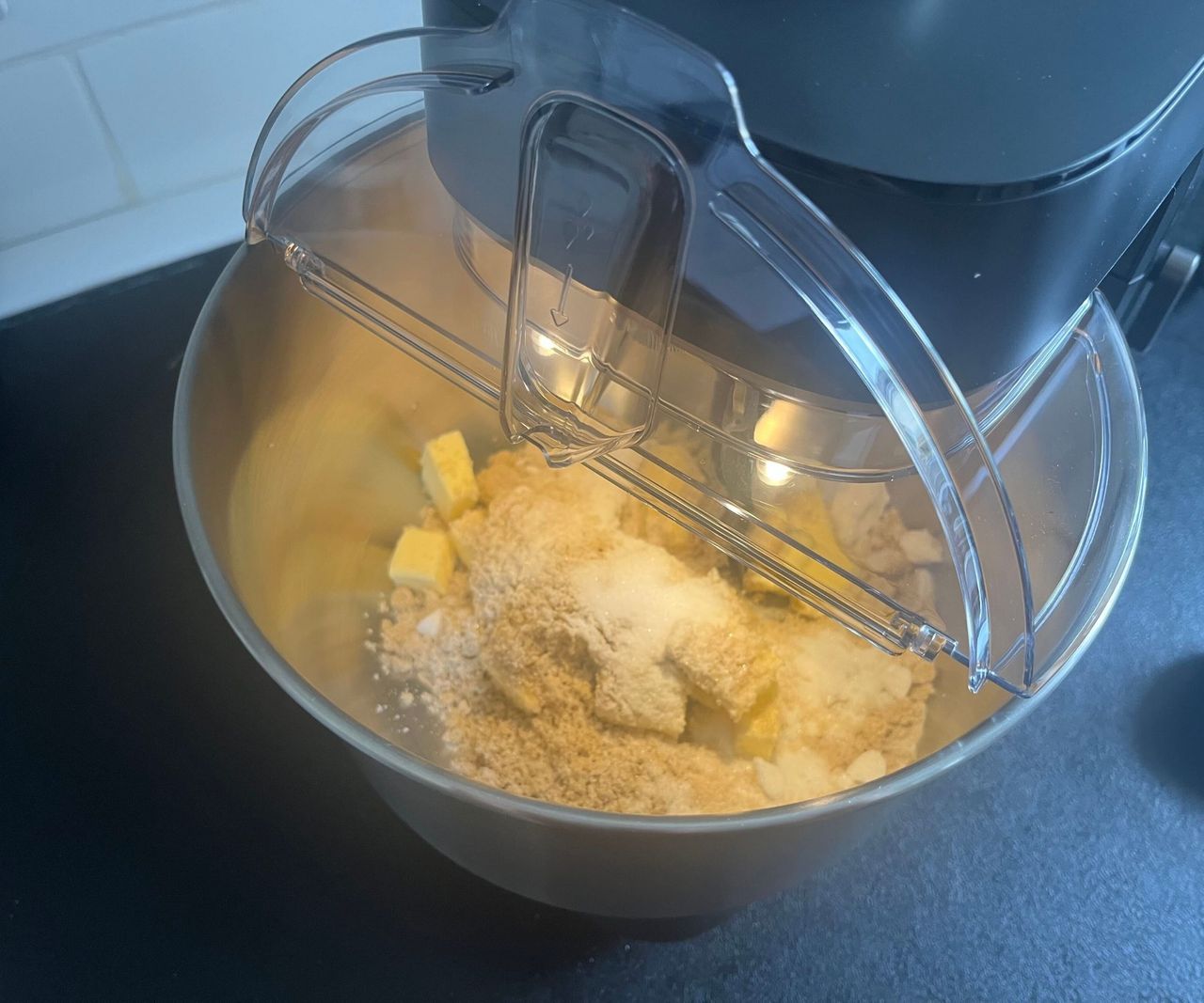 A stand mixer with a clear plastic spill lid on top, flipped open to see inside the bowl
