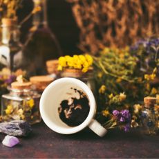 Composition of flipped over white cup with coffee grounds, colorful stones, bottles with dried flowers, oil and tincture, toy glass ball, dry herbs and a net.