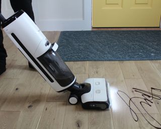 Tineco Floor One S7 steam wet-dry vacuum cleaner cleaning chocolate sauce from hardwood flooring. Yellow floor and blue doormat in background