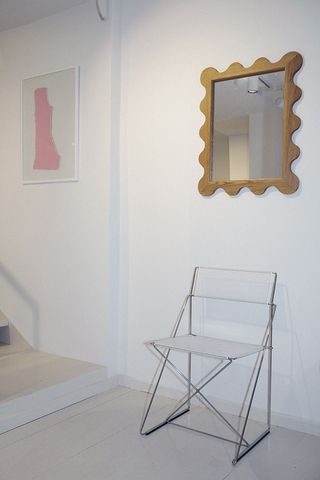A white chair on white floor. Over the chair is a mirror with wooden scalloped frame, and on the left is an artwork featuring a pink piece of paper on white background