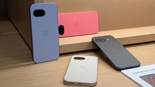 All four Google Pixel 9a phones in each color (Iris, Peony, Porcelain, and Obsidian) placed artistically against or in front of a wooden display.