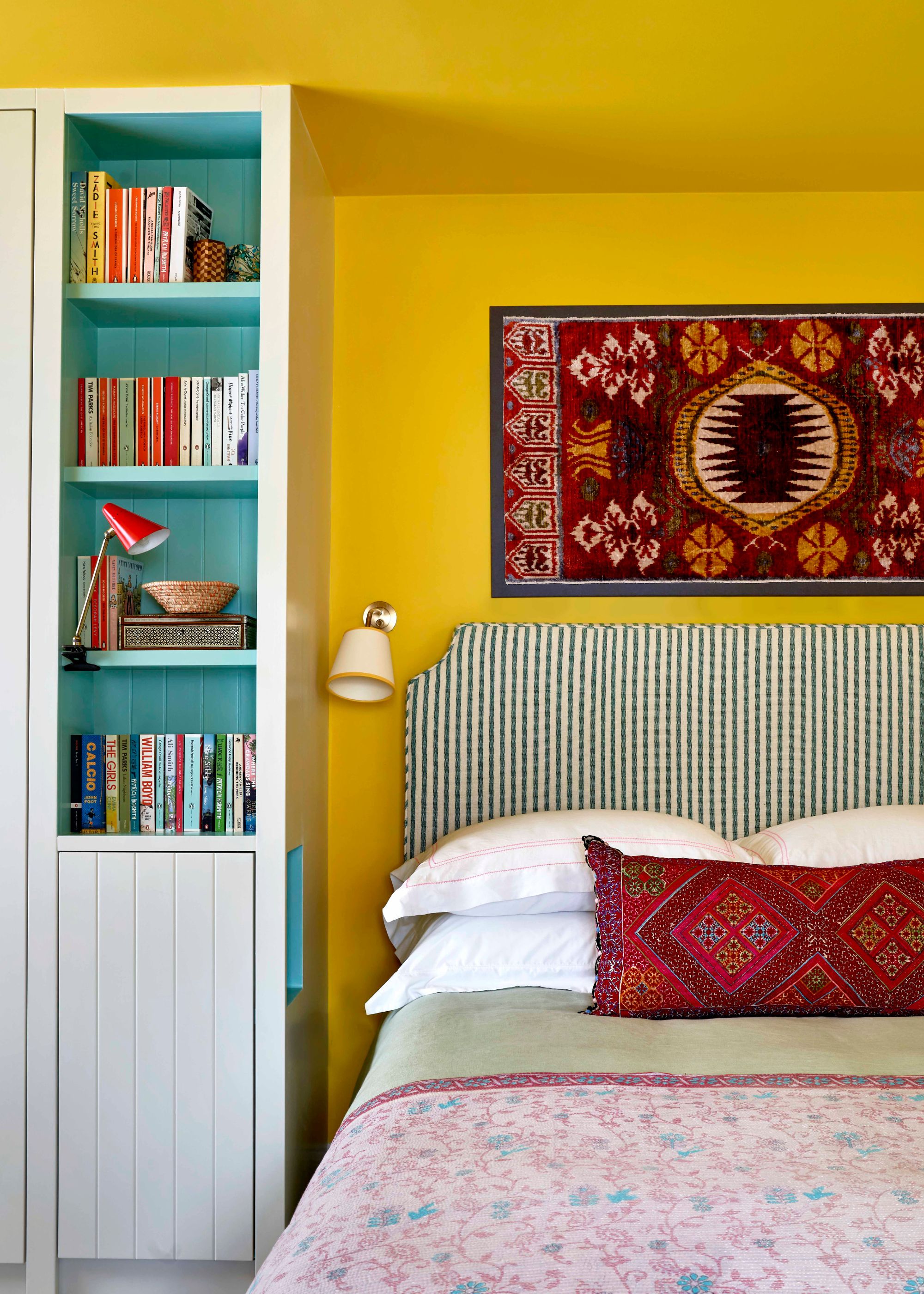 A bedroom with bright yellow walls and ceiling, a white bookcase with turquoise paint inside, a striped headboard, a red textile on the wall, and an antique red cushion on the bed.
