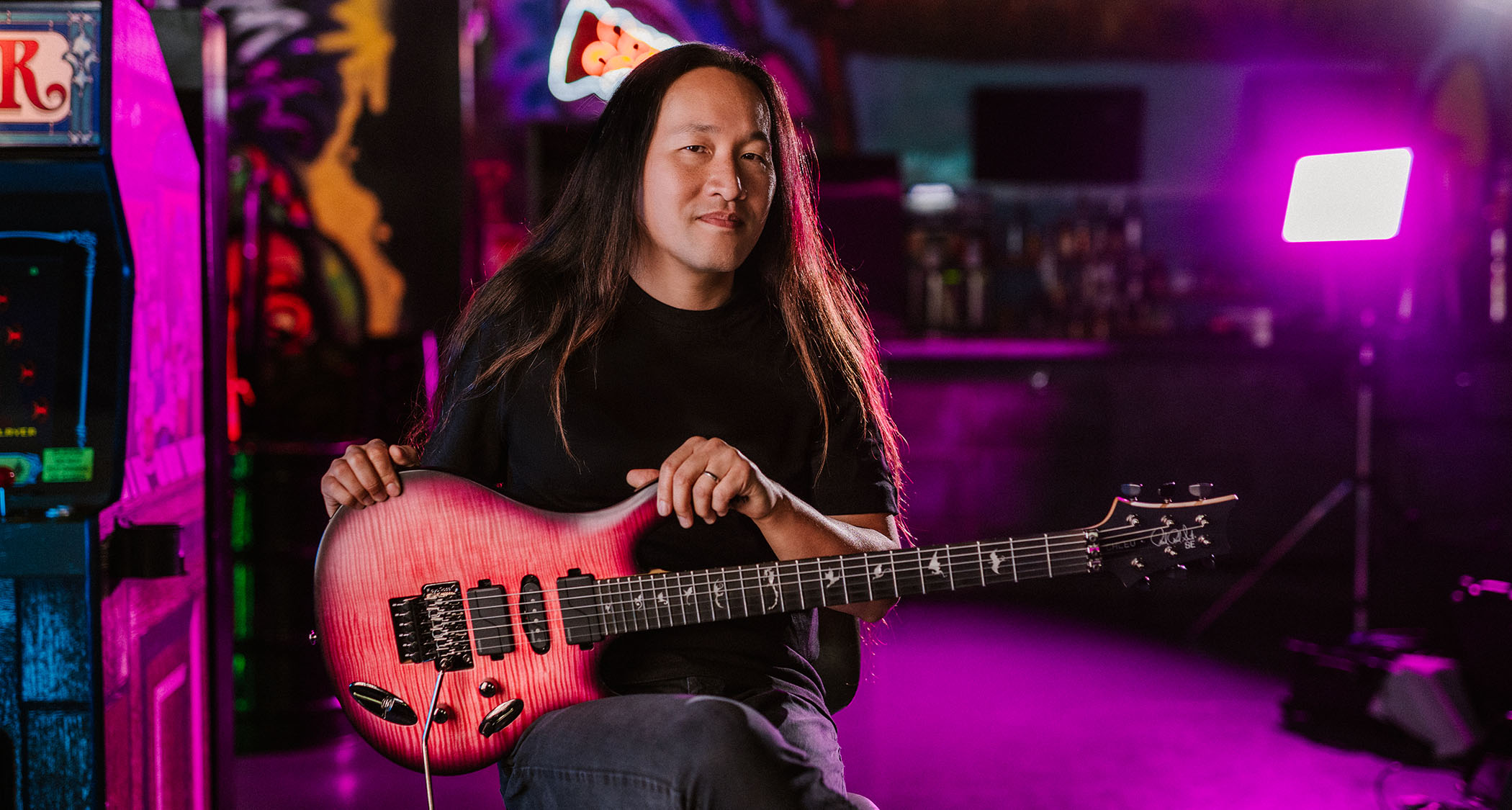 Herman Li of DragonForce sits with his new PRS SE signature model, Chleo, in a purple-lit room filled with arcade machines.