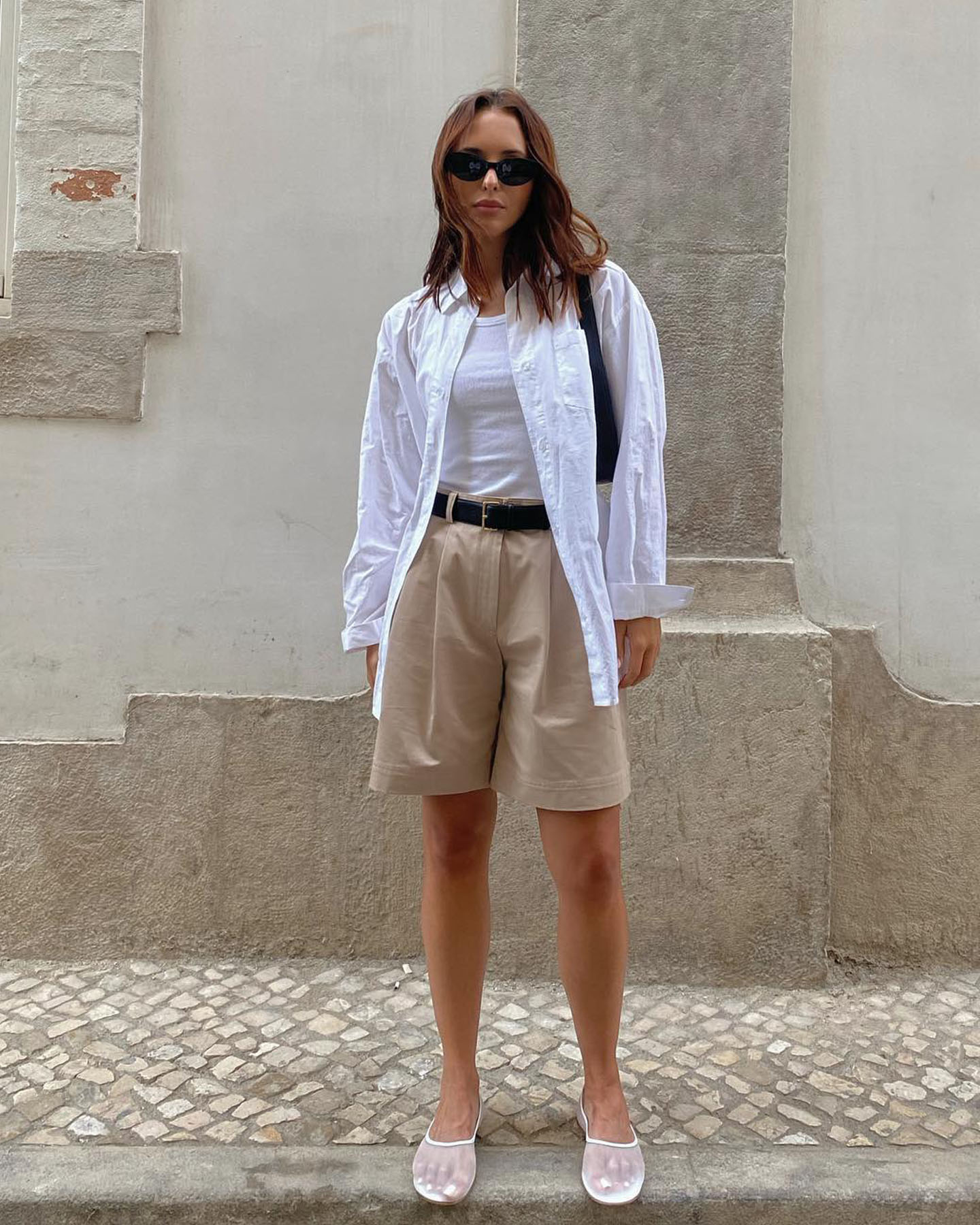 Influencer Debora Rosa wears oval sunglasses, a white button-down shirt, white tank top, black belt, khaki mid-length shorts, and white mesh flats.