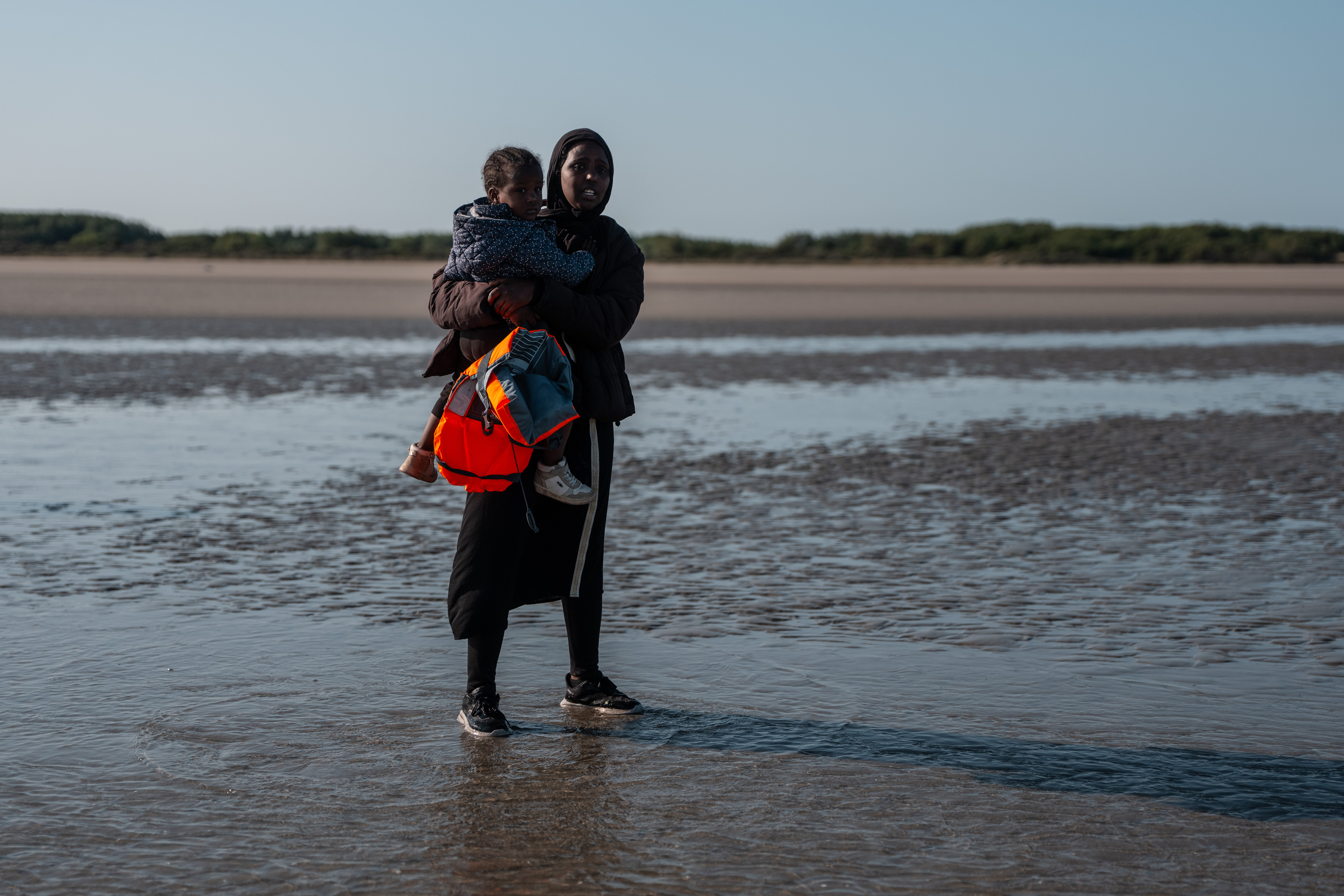 An East African woman holds a child as she waits on the beach for a migrant dinghy to sail into the English Channel on June 17, 2025 in Gravelines, France.