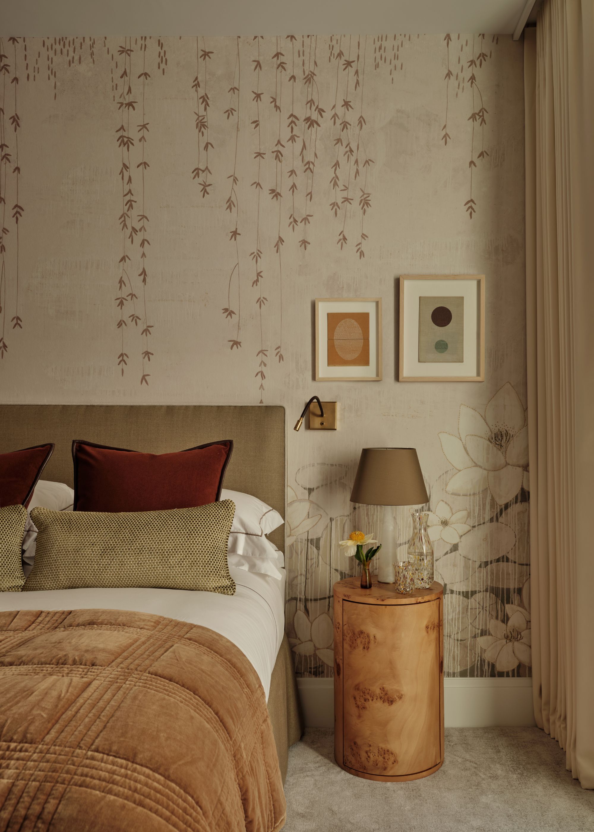 A bedroom with beige, floral-patterned wallpaper, beige carpeted floor, a taupe upholstered headboard with layered neutral bedding, and a burl wood nightstand.