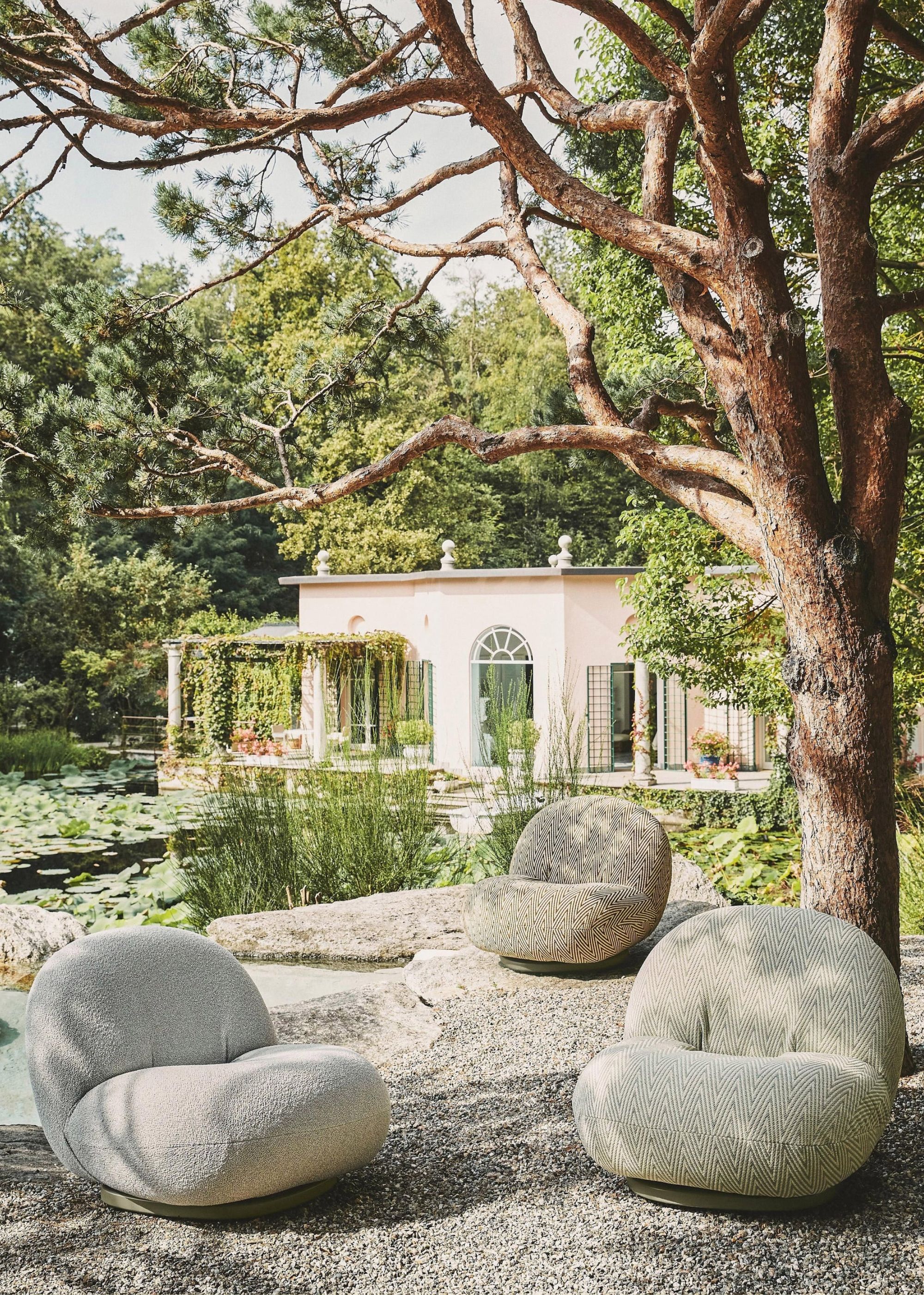 Three light colored garden sofa chairs on gravel beside a beautiful lush pond