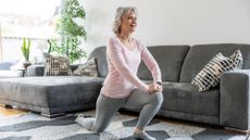 woman wearing a light pink top and grey leggings performing a deep forward lunge in a living room. there's a grey sofa behind her and grey patterned rug beneath her.