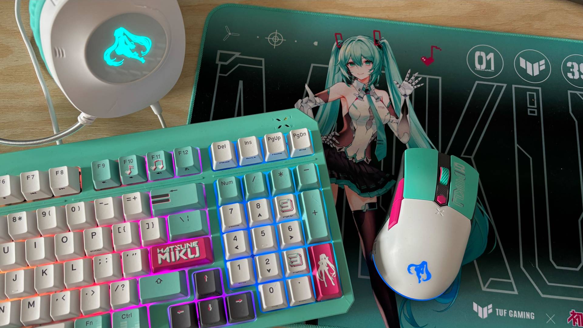 Hatsune Miku x TUF Gaming collaboration ft. Keyboard, Mini Wireless Mouse, Headset, and Mouse Mat against pale wood background