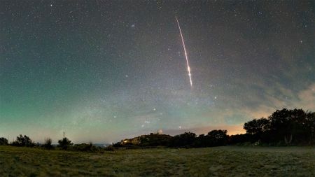 a red-tinted streak from a meteor plunges through a greenish-blue sky above the dome of an astronomical observatory