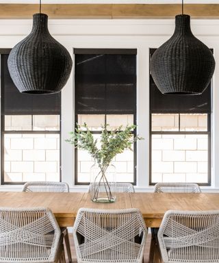 Warm wood dining table with woven string dining chairs, glass vase with greenery, and duo of large black woven pendant lights hanging over tabletop.
