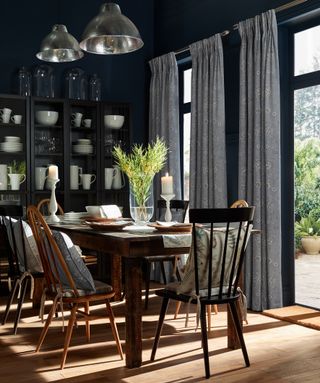 Dark dining room scheme with floor length drapes, mix and match spindle chairs, and dark glazed cabinet filled with white dinnerware.