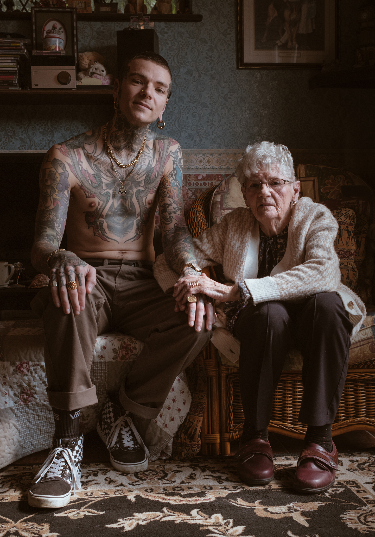 A tattooed man sits beside an elderly woman on a floral couch. They hold hands, conveying warmth and connection in a cozy, vintage room