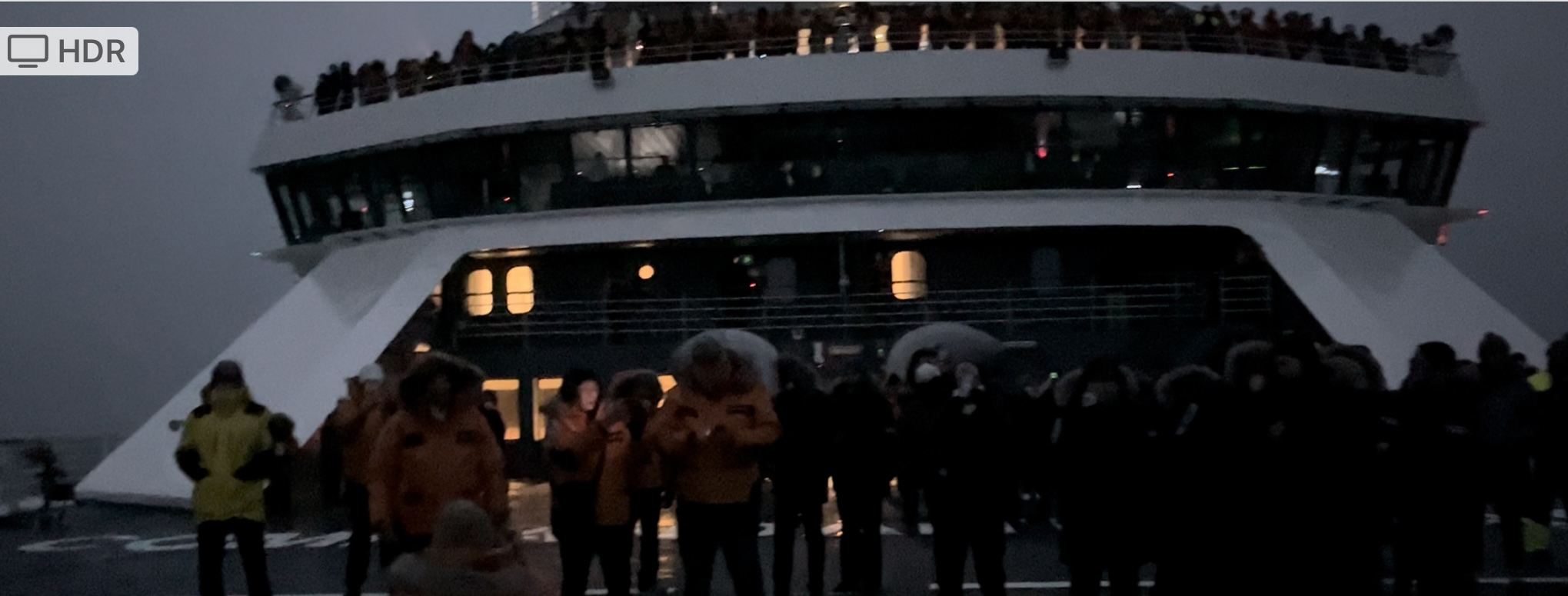 a dark image of a ship with people standing out on the deck and thick gray clouds above.