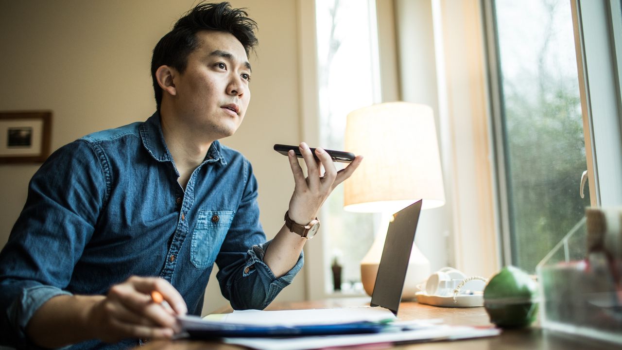 A young man sits at his desk in front of his open laptop and appears to be talking into his smartphone while it&#039;s on speaker.