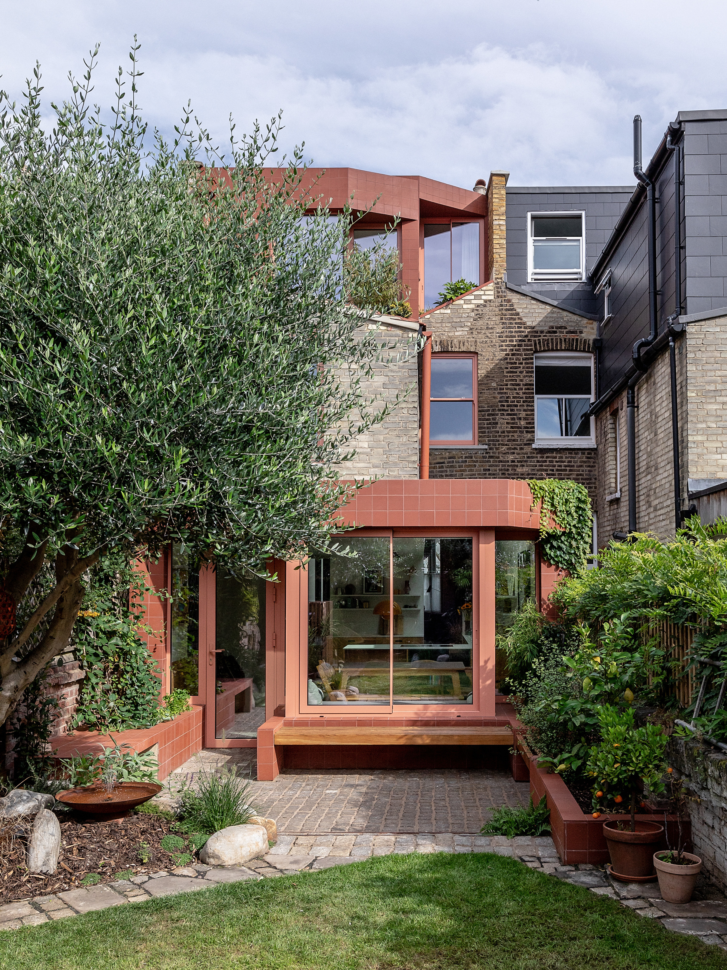 An extension with square terracotta tiles surrounding the window and door frames of a kitchen extension on the ground floor, as well as on the top floor, and the same color is brought across the window frames and guttering as well, with an abstract-shaped garden in front of the extension