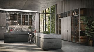 A modern designed kitchen with Belgian stone counters and a mellow grey finish, tinted glass panels and shelves in custom metal frames. 2 grey stools by the counter and a large green plant in a gold pot at the corner of the kitchen