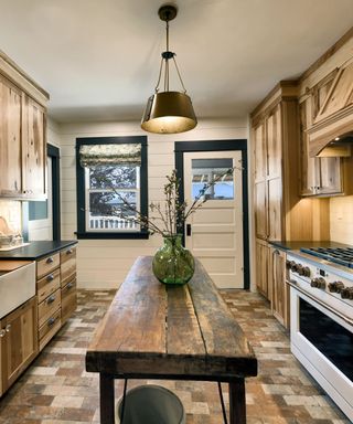 A kitchen with wooden cabinets to the left and right, white walls, and a wooden kitchen table in the middle with a green vase on it and a black pendant light hanging over it