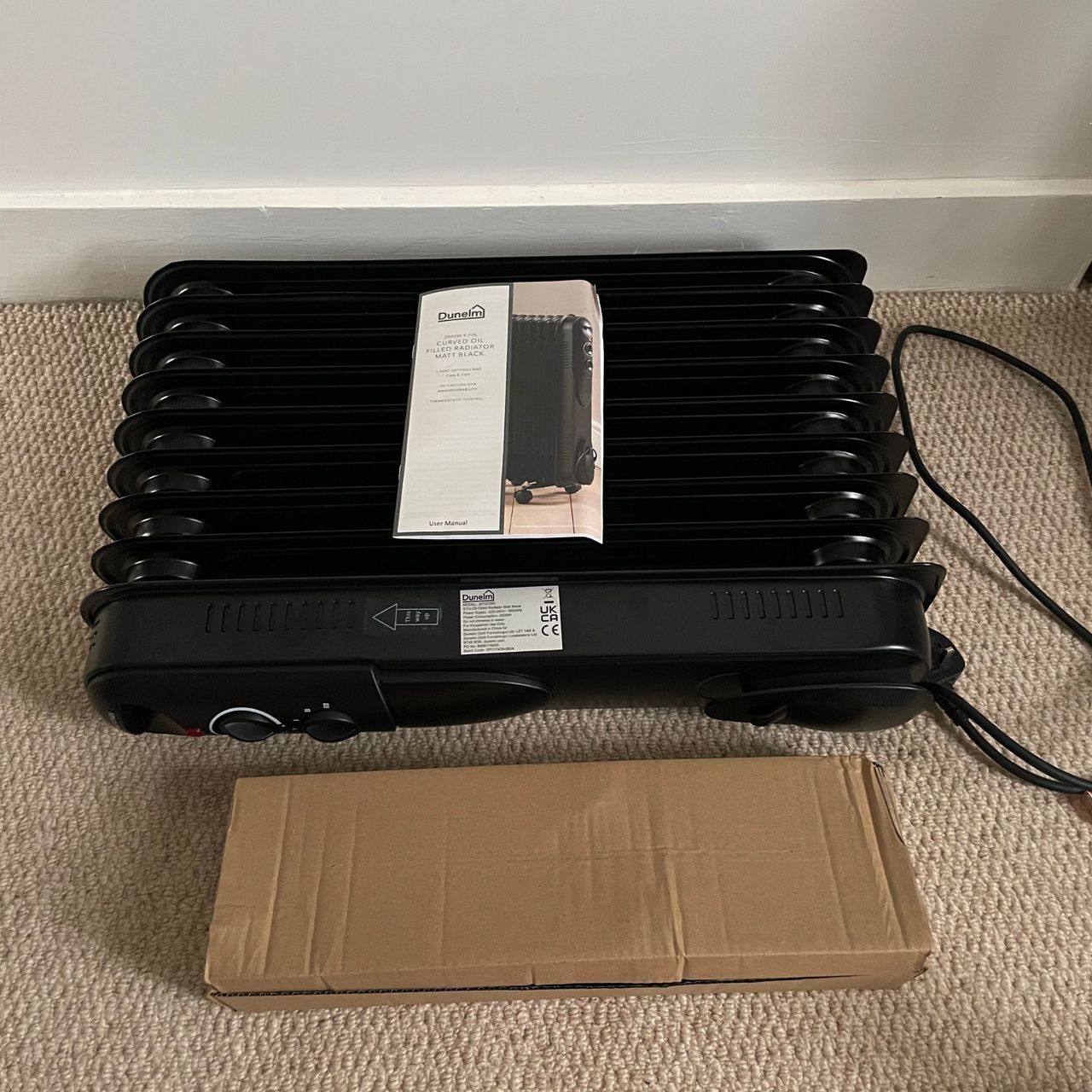 Black Dunelm oil-filled radiator laying on its side on carpet, with the instruction manual laying on top of it and a smaller box in front of it