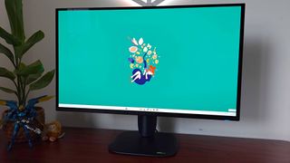 Alienware AW2725Q gaming monitor with green Kiki's Delivery Service wallpaper on Windows 11 sitting on woodgrain desk.