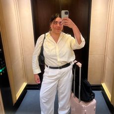 Woman taking mirror selfie in hotel elevator while traveling with carry-on suitcase.