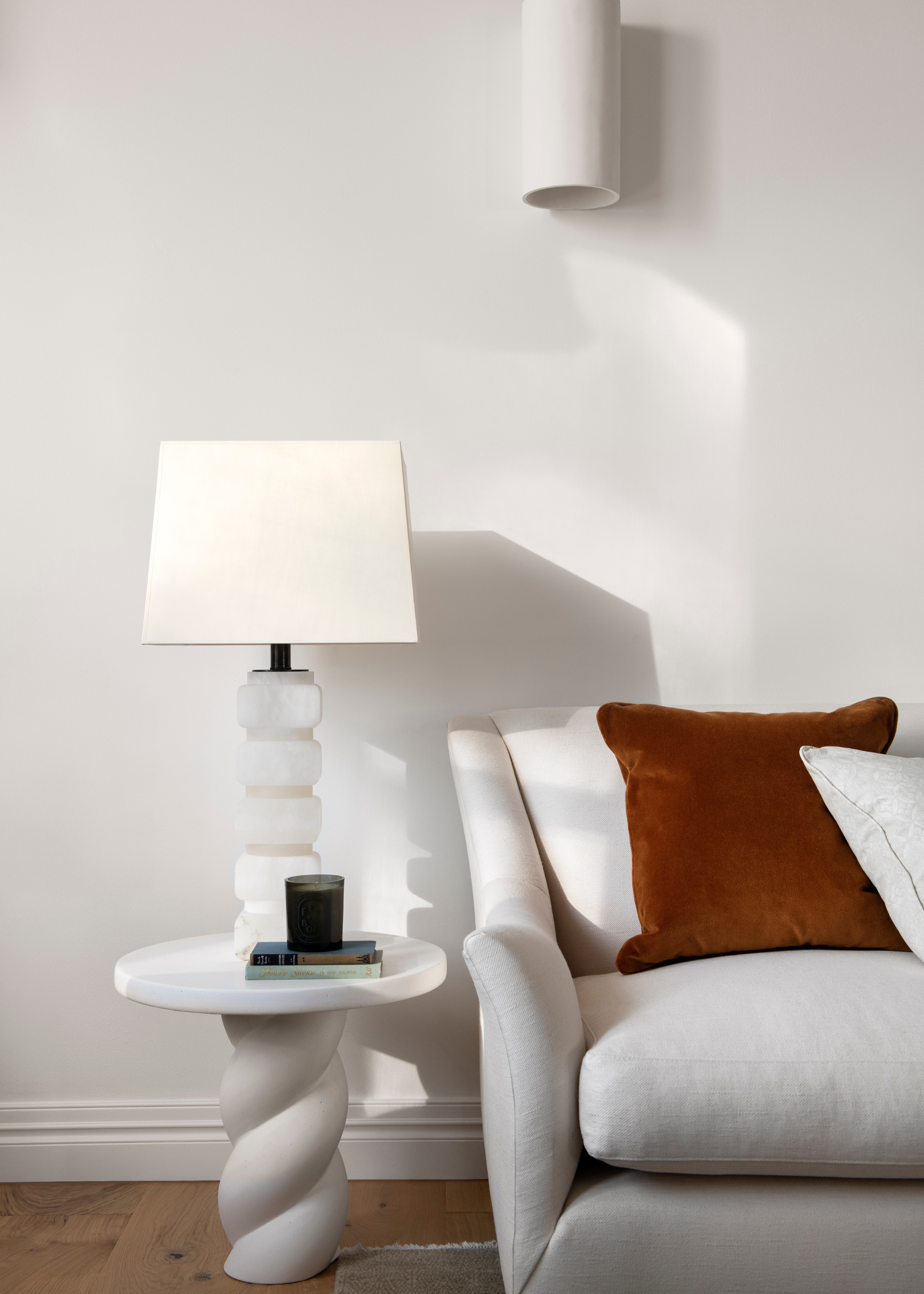 A white living room corner with a white couch, a white side table with a stacked table lamp and a scented candle