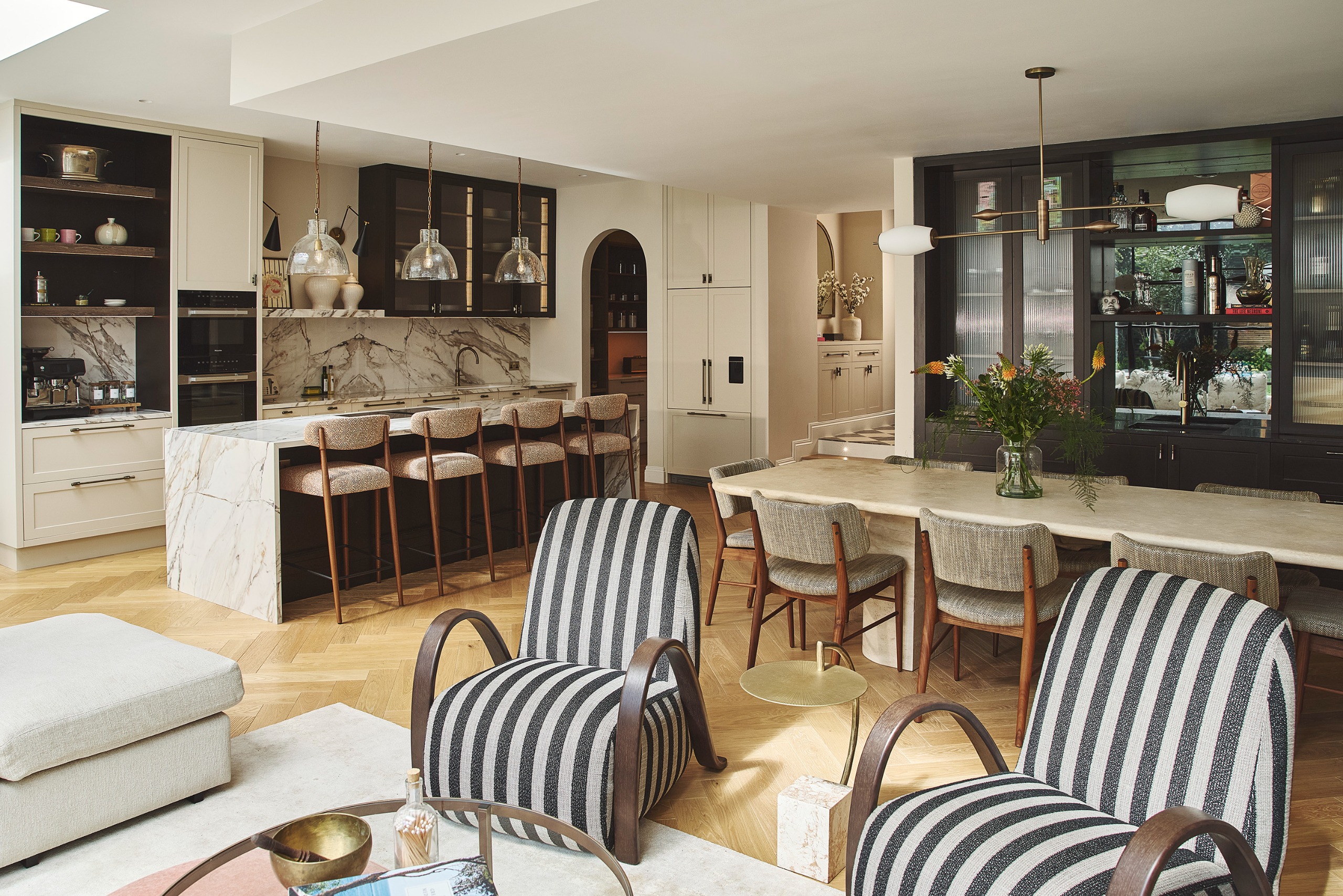 a open plan kitchen and dining room with a seating area. striped chairs are in the foreground, and an island and pantry in the rear