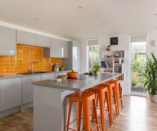 A kitchen with colour pop splash back tiles and orange bar stool seating. The cabinets and surfaces are a neutral grey