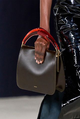 The frame bag trend is shown in a close photo from the Ferragamo fall 2023 runway show with a model wearing a black patent leather dress with a black frame handbag