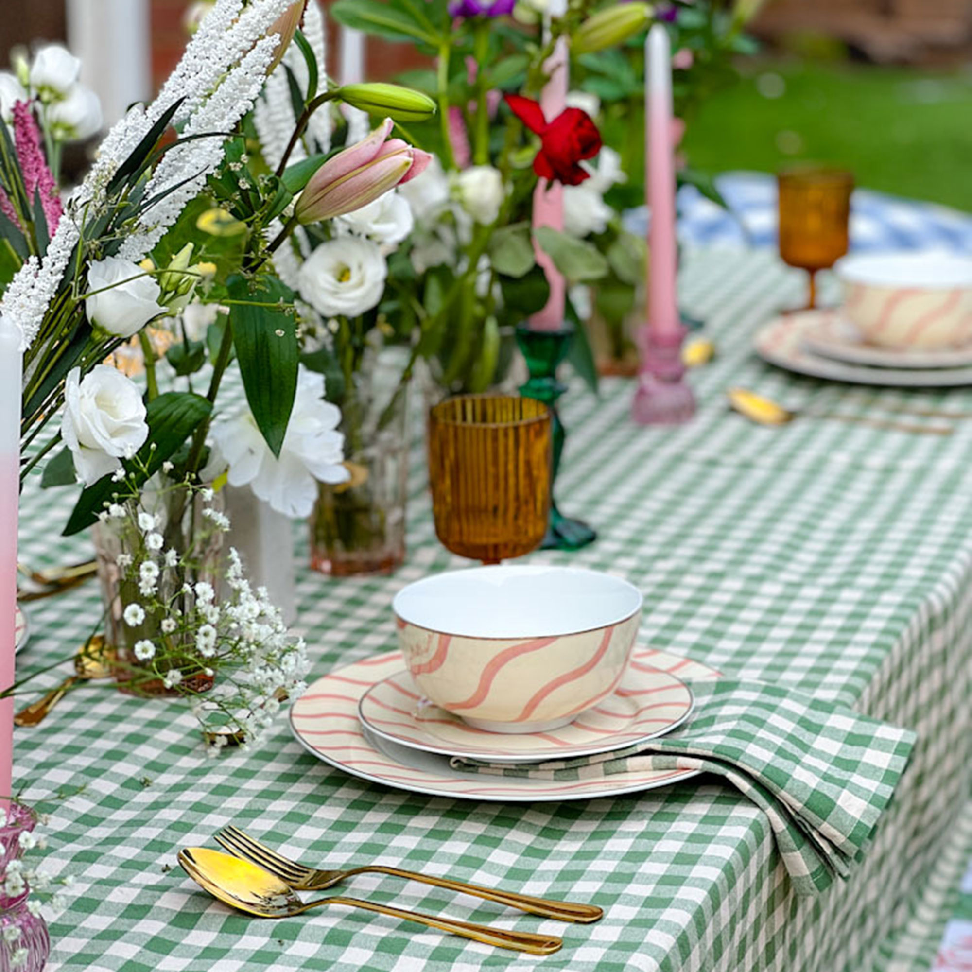 Overhead view of table decorated with green and white gingham tablecloth, flowers and foliage, gold cutlery and flatware with pink and white squiggle pattern