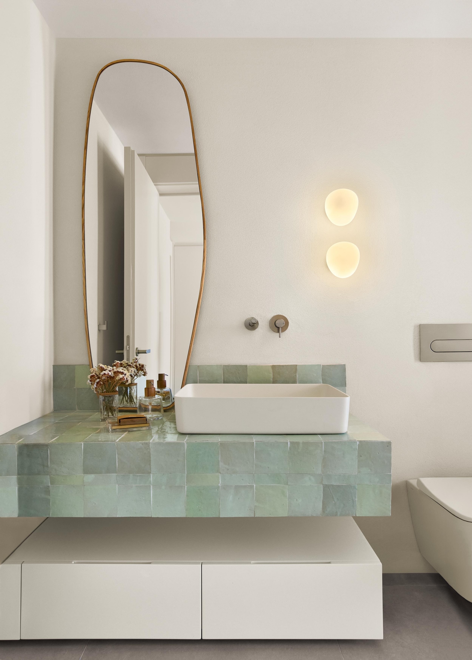 A pale green tiled vanity unit with a sit on basin, long curved mirror and frosted glass wall lights