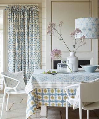 White and blue kitchen with matching curtains, tablecloth and cushions, white dining chairs, table decorated with vases & kitchenware