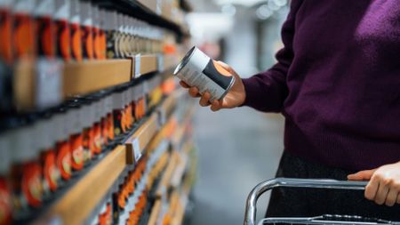 Woman holding a can of soup and looking at the label in the supermarket, holding basket, representing a list of ultra-processed foods