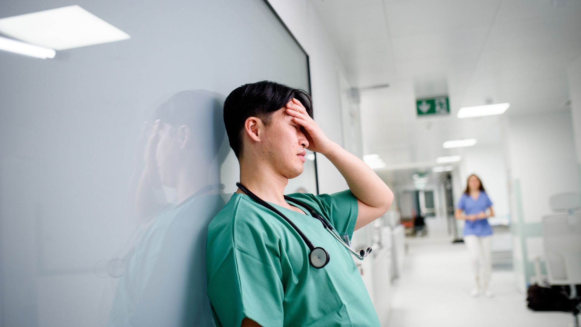A male nurse wearing green scrubs with a stethoscope around his neck, leaning against a wall with his hand on his head visibly tired.