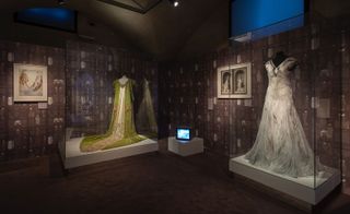 A display of 2 outfits enclosed in a glass box with white platform base in an exhibition room with brown floors and walls. on the left: Green court cape in silk velvet with embroidery; on the right: a dirt white gothic style short sleeved bridal dresss