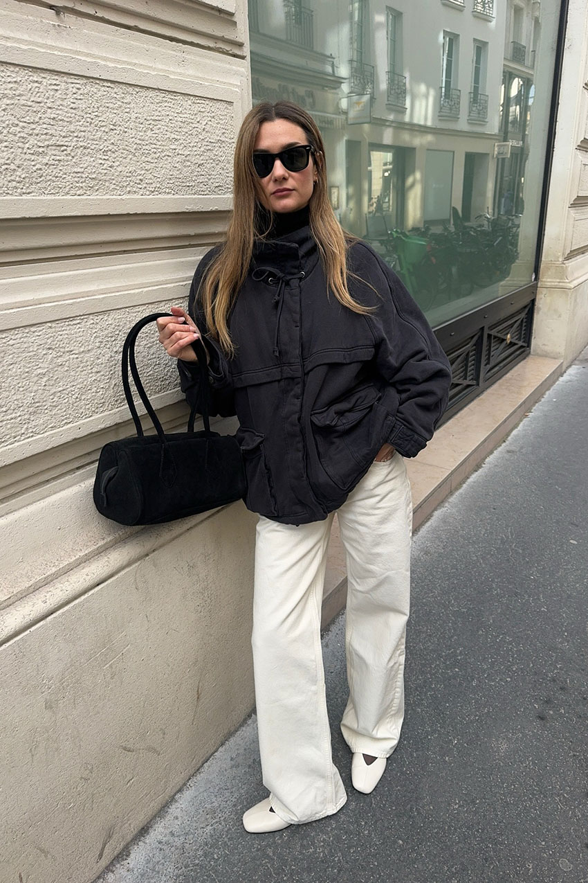 a woman wearing a black jacket, white pants, white flats, and a black shoulder bag