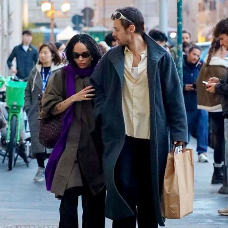 Zoe Kravitz walks down the street holding hands with Harry Styles. She wears a khaki trench coat with a purple scarf and patent shoes, whilst he wears a blue trend with a yellow shirt.