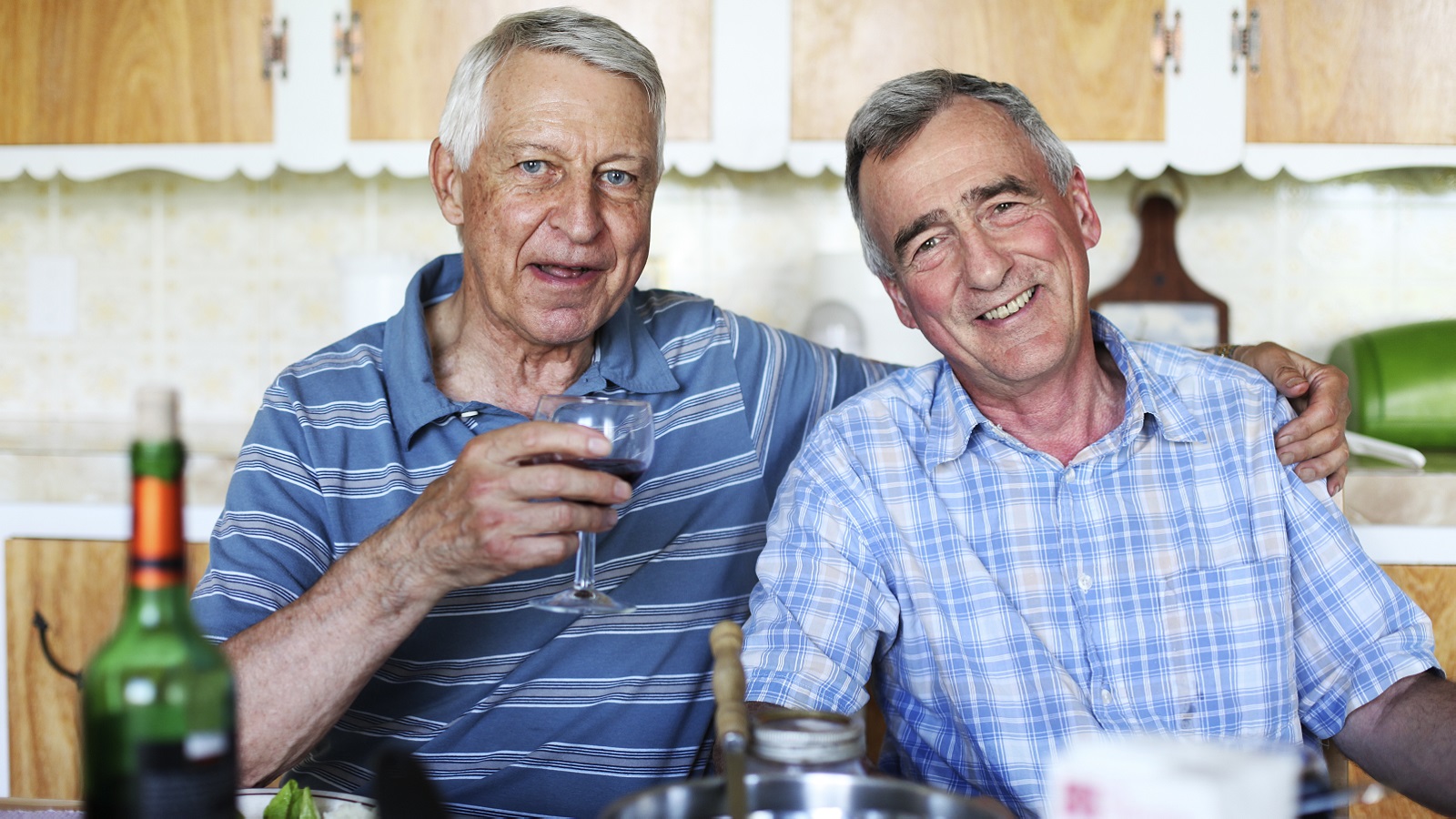 A gay male couple sit at the kitchen table enjoying a bottle of wine.