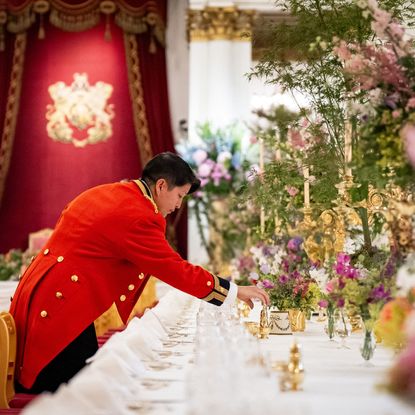 A red uniformed palace employee setting an elaborate table with candles and flowers in front of a red throne