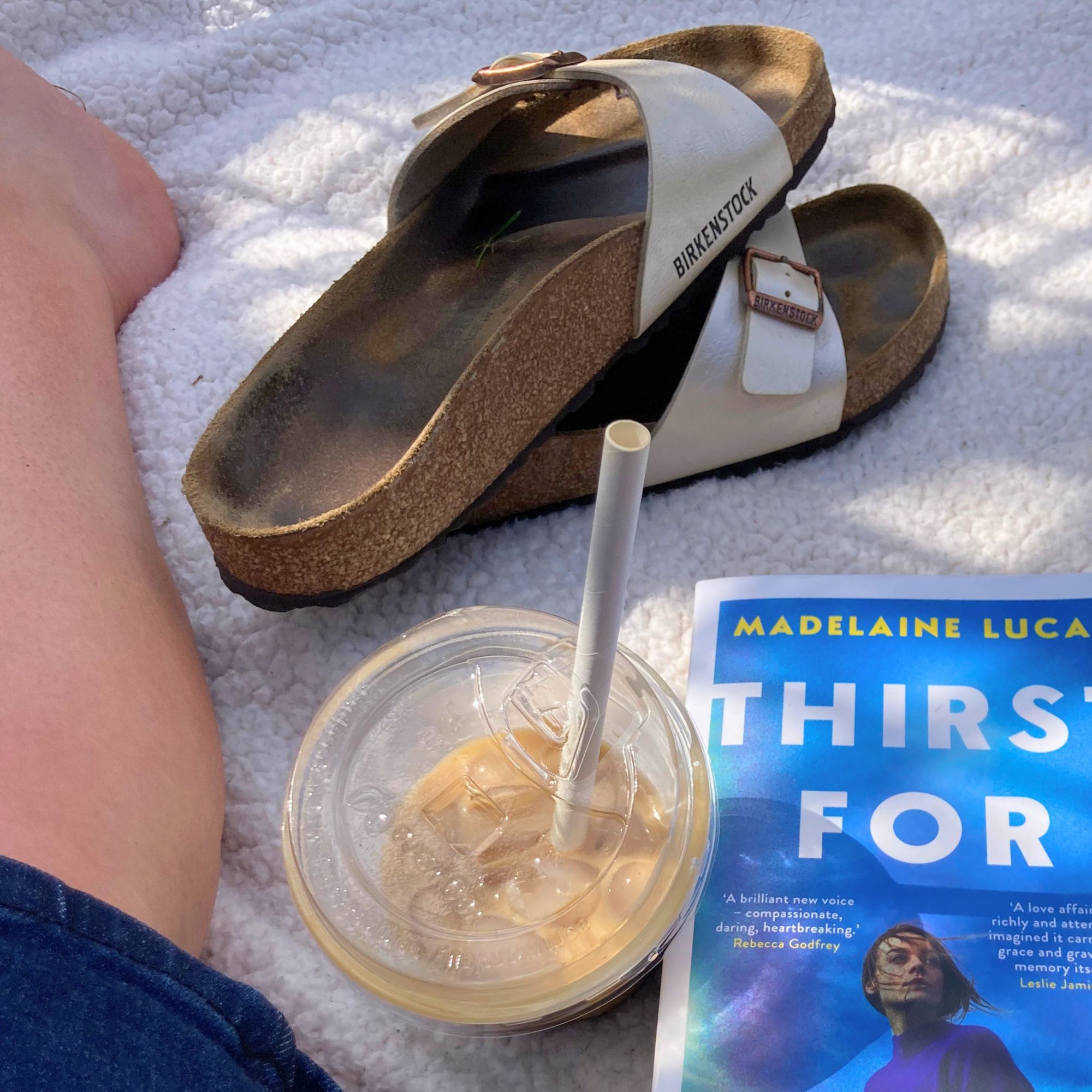 A real-life shot of writer Heidi Scrimgeour's Birkenstock Madrid sandals pictured on a picnic blanket beside an iced coffee and a paperback book with her legs visible in the background