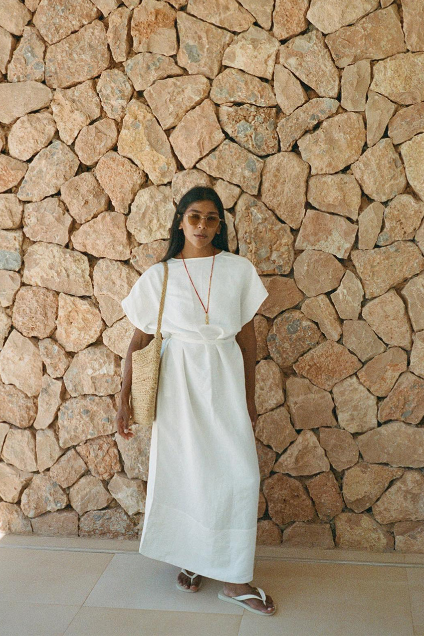 A woman standing outside against a rock wall wearing a white linen maxi dress with white flip flops, a raffia bag, aviator sunglasses, and a red lariat necklace.