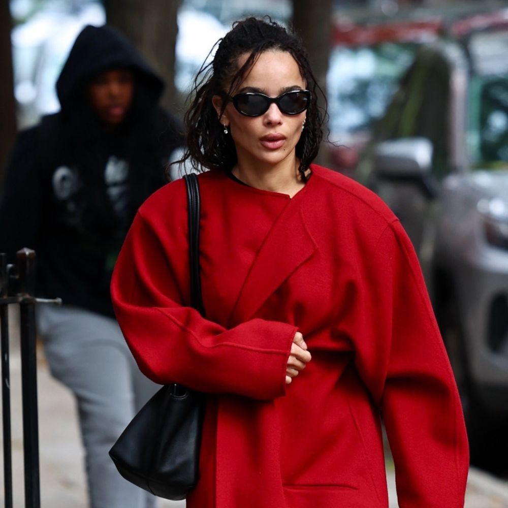 Zoe Kravitz walks down the street in New York wearing a red collarless coat with black trainers, a black leather bag and black sunglasses. She holds a transparent shopping bag and a brown paper bag.